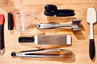 A top view of various Oxo kitchen utensils laid out on a kitchen counter, including: can opener, meat tenderizer, microplane zester, tongs, measuring cup, silicone brush, and silicone spatulas.