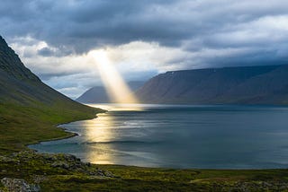 Light streams through clouds onto a large body of water surrounded by hills.