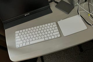 A wireless Apple Magic Keyboard and a white Magic Trackpad are placed side by side on a light-colored desk. A KYY portable monitor, a connected external hard drive, and several tangled cables are also visible