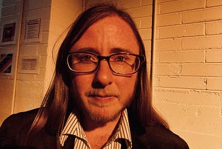 The author is standing in front of a white brick wall wearing a black suit and a pinstriped shirt. He’s wearing glasses and a serious face. He has long brown hair, a beard and a mustache.