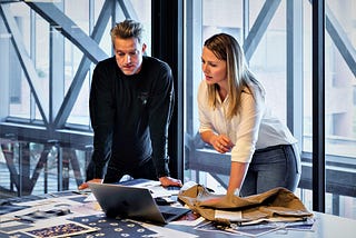 man and woman in workplace looking at computer on desk with glass wall behind them