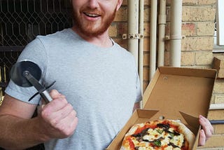 A photo of the author in a grey T-shirt, a small pizza in his left hand and pizza slicer in his right hand. He’s standing next to a small wooden-slatted table, and in front of a mesh-screen door and a tan brick wall with pipes running vertically and horizontally along it. He has an orange beard and short, light orange hair.