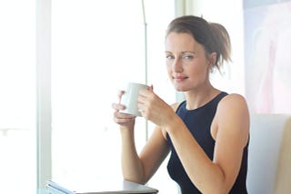 woman smiling as she holds a cup of coffee while pausing at her desk