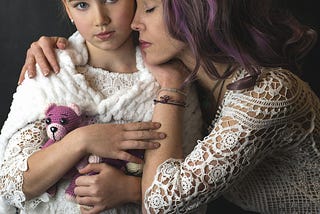 Mother and daughter, both dressed in white lace. The girl has small purple stuffed bear and a purple hair tie, the mother has purple highlights in her hair.