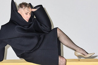 Alan Cumming in a black robe and high heels, posing suggestively for the camera
