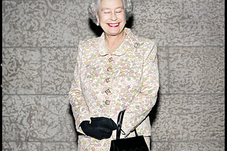 The late Queen Elizabeth the second standing in the center of the frame. She’s wearing a yellow floral print jacket and skirt. Wearing black gloves with a black purse hanging from her left wrist. Her eyes are closed and she’s smiling with lips painted red. It’s not a regular boring image of her because of the look on her face. Photograph by Christopher Wahl.