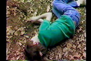 A Boy in a green shirt and blue pants and brown hair lies in some leaves.