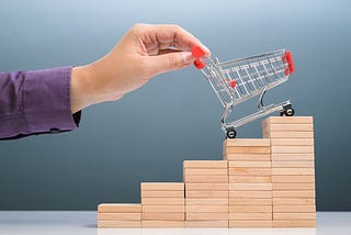 A person’s hand pushing a small shopping cart up a set of stacked blocks arranged like a staircase