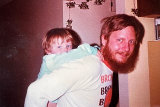 My dad and I, approximately 1984. Description: an older photograph of a white man with red hair and a longish beard with mustache and blue eyes looks at the camera and smiles. Me, a small white toddler of about 3, hangs on for a piggyback ride. I’m wearing a light blue shirt with long sleeves.