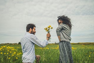The picture shows a man and a woman in a beautiful green field. The man is down on one knee and has picked some yellow flowers from the field. He is holding them out to the woman. She is looking at him shyly. It’s unclear whether she will accept them or not.