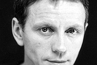 A black and white headshot of Brent Carver. He is looking directly into the camera, slightly smiling.