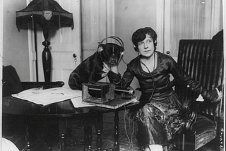 Black and white photo of Mme. Asta Souvrina, “the Bernhardt of Russia,” sitting beside a dog named Buster. Both are wearing headphones, listening to a radiophone device on a table in front of them. Taken around 1922 by Underwood & Underwood.