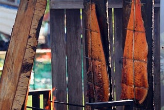 Color photo of large pieces of salmon being smoked outside in a makeshift smokehouse on a rack.