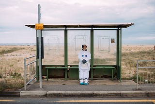 An astronaut waits at a glass bus station alone on the plains.