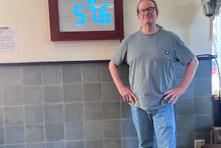 Me standing in front of the clock I built for UQBAR Café in Berkeley, CA. The clock is mounted on the wall behind me and measures 36 inches tall by 29 inches wide. The front panel shows three rows of numbers: “5:07”, “7131” and “57.6”.