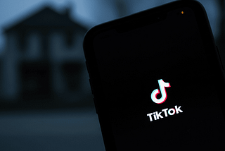 TikTok logo in pink-white-blue glow on smartphone screen at night, with blurred house in dark background.