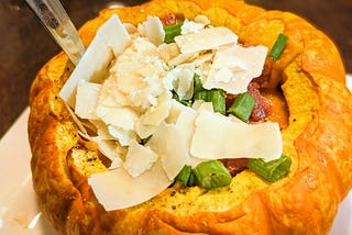 A roast pumpkin used as a bowl for pumpkin soup, topped with shaved parmesan, green onions, and crumbled bacon.