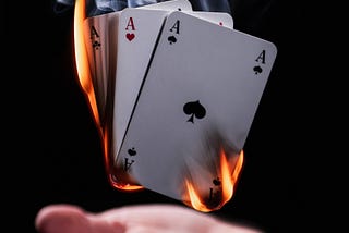 A white hand outstretched with playing cards hovering above it. The cards are on fire.