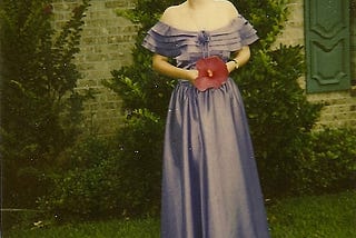 A redhead girl with her hair up wears a long, lavender, off-the-shoulder dress and holds a large red hibiscus flower in front of a huge bush in front of a ranch-style house.