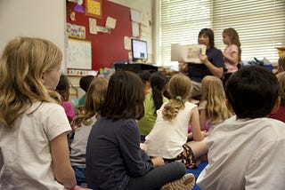 Children in a classroom being read to by a teacher.