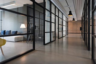 Image of office featuring hallway with glass doors leadign to meeting rooms. Meeting room on the left of the picture with couch and yellow chair.