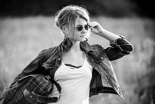 A black and white image of a young woman wearing sunglasses, holding a bag.