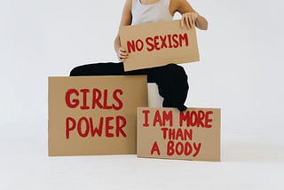 Image of a person with signs stating: “no sexism”, “girls power”, and “I am more than a body”.