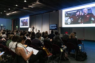 Man on stage between two screens showing a Star Trek TNG image of Picard and Riker conferring while Data looks on.There’s a screen of captions on his left. Seated audience in the foreground of shot.