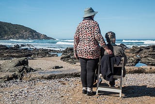 Color photo of two older women by a lakeside, on a clear day with blue sky. One is seated, the other standing, with one hand holding unto the back of the chair; as they both look out at the view.