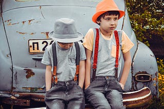 Two boys wearing gray shirts sitting on gray vehicle bumper