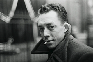 Black-and-white portrait of Albert Camus, a man in his thirties with combed-back dark hair, glancing over his shoulder toward the camera. He wears a heavy overcoat with the collar turned up and a lit cigarette between his lips, against a blurred, dimly lit background.
