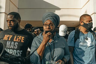 A Black woman speaks at a rally with two Black men on either side of her. Several other Black people stand behind the trio.