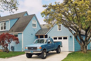 Picture of a cookie-cutter blue home in a lower middle class neighborhood with a pick up truck in front of it; the american dream encapsulatyed in an image.