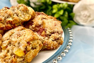 scones studded with peach and raspberry on a white plate on a clear charger with silver ball rim. the plate sits on a soft white background with a plate of scones surrounded by white flours and greenery. Inspired by Penelope Featherington