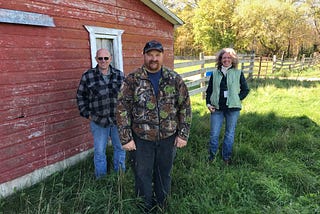 three people stand in front of a building with a wooden fence and trees in the background