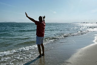man standing at the sandy beach with the waves crashing into his feet and he rise his hand as if he embrance the morning sunny day and blue sky