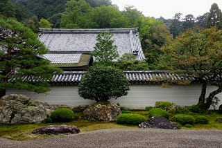 Green trees behind a rock garden, in a zen garden. In the background is a wall with Japanese style shingles.