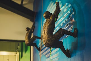 Two bronze figurine men climbing a wall with binary code printed on it.