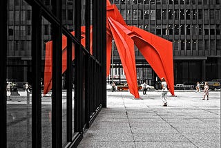 Half of a large red metal sculpture appears around the corner of a modern skyscraper, creating an illusion of symmetry, with that part of the red metal sculpture reflected in the building’s windows. A handful of relatively small people walk around in the middle distance.