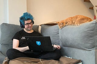 A person in a black t-shirt on a blue couch shows the screen of the laptop he is holding to an orange cat perched on one of the pillows, as if it is can read the screen.