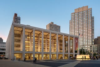 picture of a David Geffen Hall, a building with tall, thin rectangular columns marking the exterior of the building, which lets a viewer see the interior of the building through windows that extend from the ground to the roof. It is sunset and the windows are illuminated with gold light. A red New York Philharmonic banner hangs vertically between the two columns all the way to the far right.