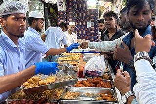 At a snacks stall, wide range of snacks are placed between the customers and the servers. One of the servers is offering a customer with a snack.