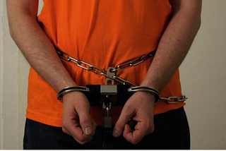 A man handcuffed man in an orange shirt and black bottoms