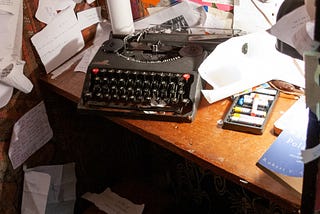 An antique typewriter sits on a desk in a corner. Papers are strewn about the desk and messily tacked to the walls over the desk.