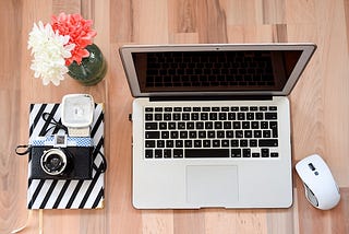 In this picture: an open laptop on a wooden desk next to a wireless mouse, a small flowerpot, and a striped, black and white planner on top of which is a camera a flash.