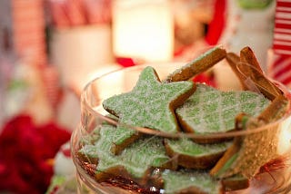 This is an image of Christmas cookies. It’s a close-up of a glass jar with star-shaped cookies with green and white icing. In the background, blurred, are red and white objects and a lamp, making for a nice overall Christmas-feel with red, white and green colours.