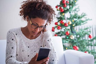 Black woman looking down at her phone with a concerned look on her face, with a Christmas tree in the background.