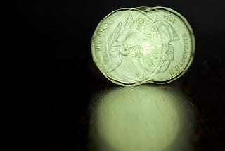Image of a spinning Canadian Loonie coin, you can see both sides of the coin in the image, black background, dark wooden surface with a diffuse reflection of the spinning coin. Image courtesy of Jamie McCaffrey via Flickr & Creative Commons Licensing