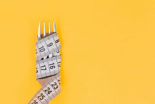 A bright yellow background with a fork wrapped with a cloth tape measure as if it were spaghetti, indicating the prioritization of body size over eating.