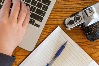 Top down view of laptop, camera, and notebook with pen